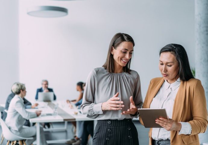 Shot of two female coworkers having a discussion in modern office. Businesswomen in meeting using digital tablet and discussing business strategy. Confident business people working together in the office. Corporate business persons discussing new project and sharing ideas in the workplace. Successful businesswomen standing with their colleagues working in background.