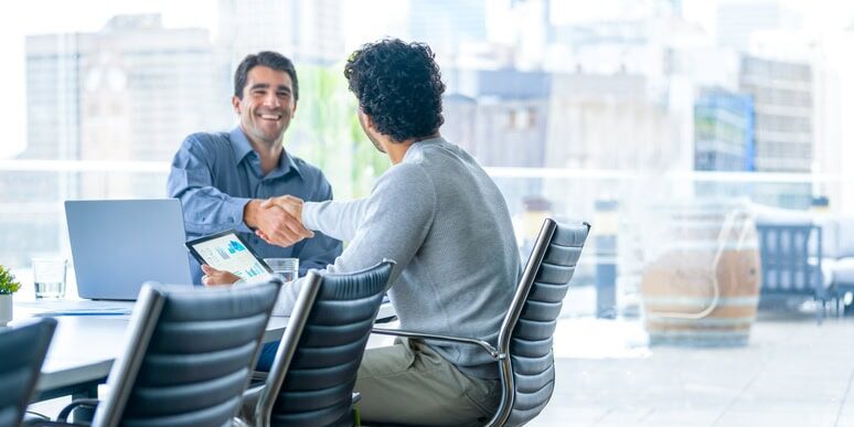 Two businessmen working on a digital tablet and laptop computer in the office in a board room or meeting room. They are shaking hands closing the deal and  a large window behind them